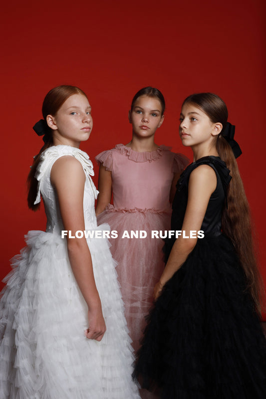 Three young girls in a studio setting against a red backdrop, each wearing luxurious, ruffled dresses in white, pink, and black with coordinating hair ribbons.