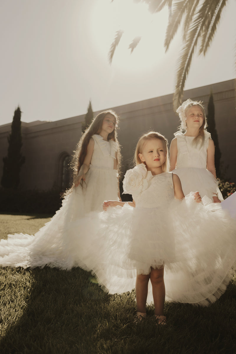 A luxurious boutique setting displaying elegant children's dresses in various colors on child-sized mannequins, surrounded by floral arrangements.