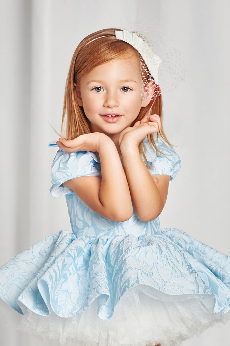 A light blue flower girl dress on a mannequin surrounded by luxurious children's dresses in shades of pink, coral, and white, set in an elegant, vintage-inspired room.