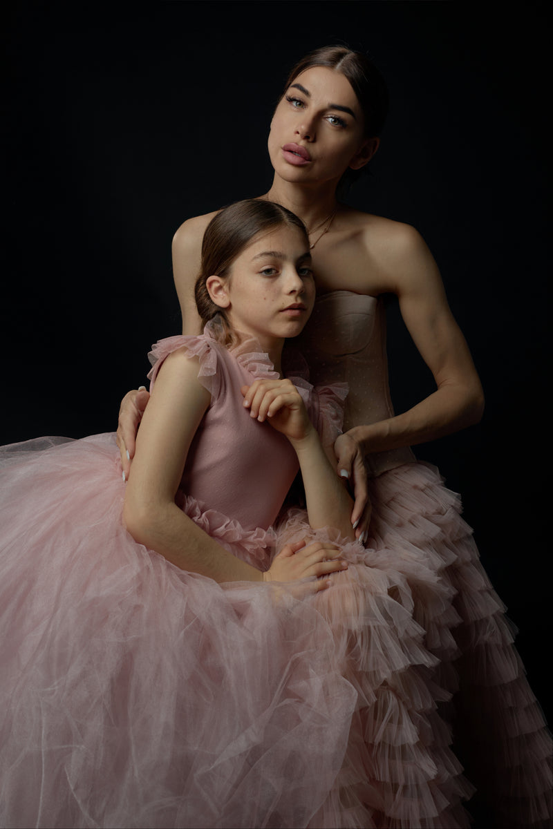 A mother and daughter in matching luxurious dresses stand elegantly in a romantic, opulent setting.