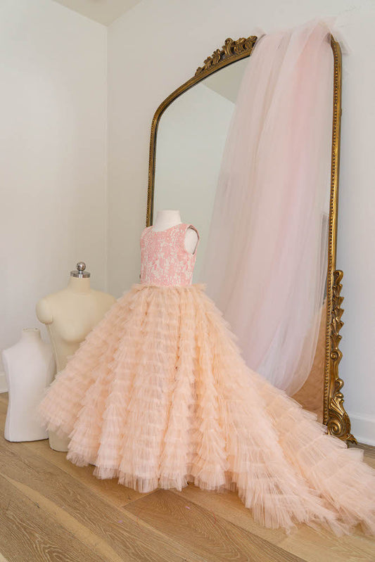 Peach layered ball gown flower girl dress with long tulle train displayed on mannequin in front of mirror