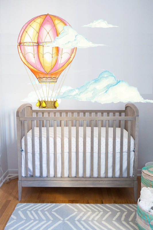 Pink hot air balloon wall decal with watercolor clouds above crib in baby nursery room